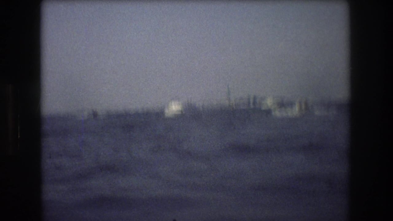 Soft 1976 New York City home-movie frame showing rough harbor water with indistinct distant vessels or shoreline forms under a hazy sky.