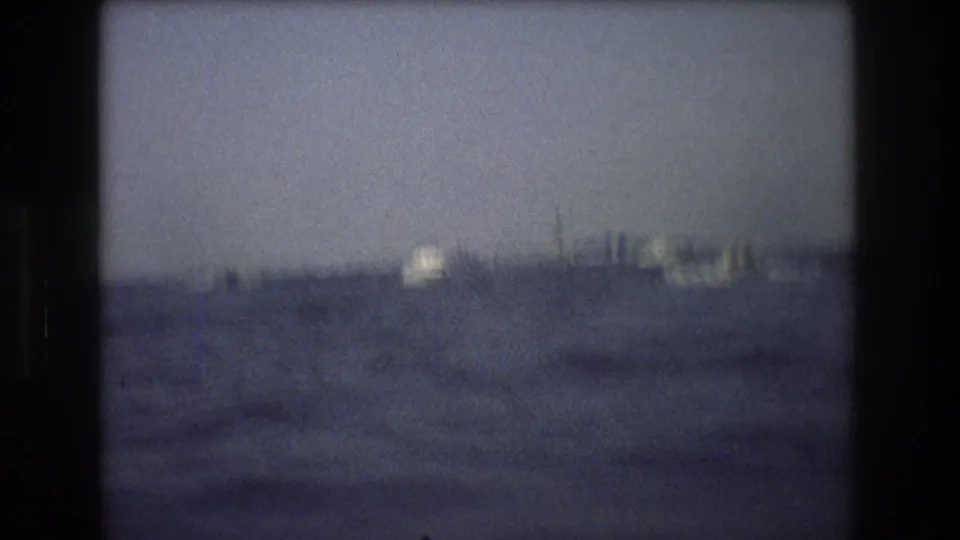 Soft 1976 New York City home-movie frame showing rough harbor water with indistinct distant vessels or shoreline forms under a hazy sky.