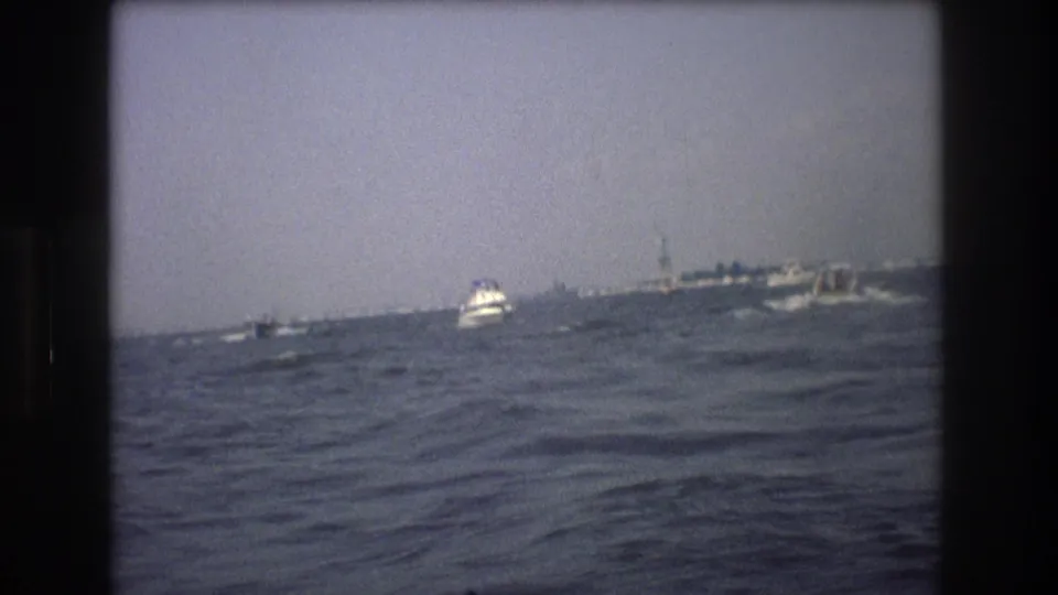 1976 New York City maritime frame with a small white boat crossing rough water and a larger distant vessel or harbor structure near the horizon.