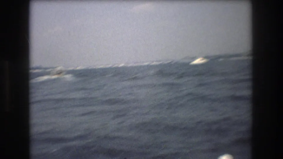 Home-movie view of rolling blue-gray harbor water with distant white wakes and indistinct boats near the horizon.