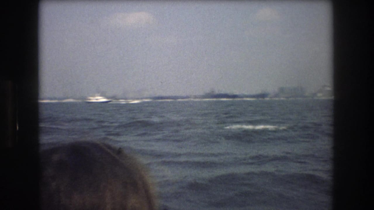 1976 8mm home-movie frame from a New York City boat tour showing choppy water, hazy distant shoreline buildings, and a partial passenger head in the foreground.