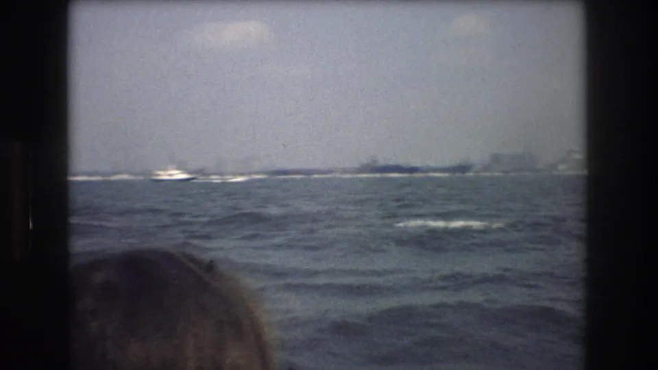 1976 8mm home-movie frame from a New York City boat tour showing choppy water, hazy distant shoreline buildings, and a partial passenger head in the foreground.