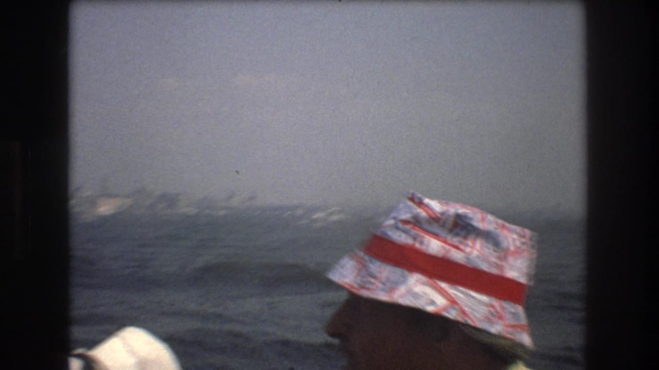 1976 New York City boat-tour frame showing a foreground passenger in a patterned bucket hat with a red band facing the harbor and distant skyline.