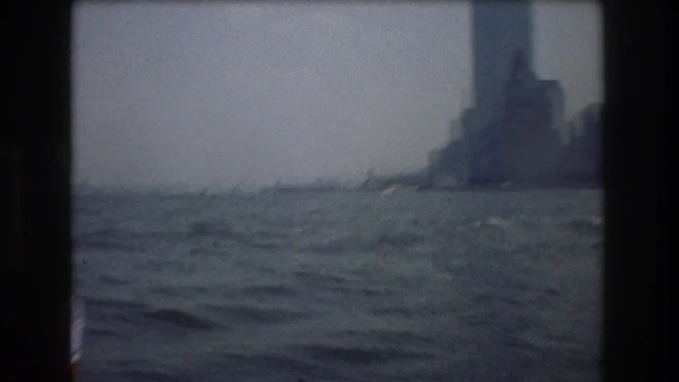 Soft 8mm frame from a 1976 New York City boat tour showing choppy harbor water and a hazy cluster of waterfront buildings in the distance.