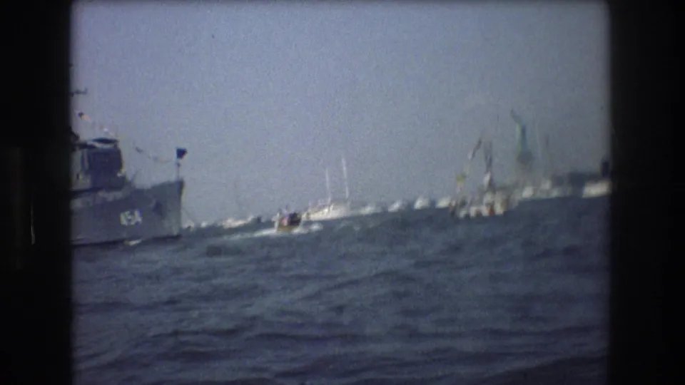 A numbered harbor vessel passes near sailboats on choppy New York Harbor water, filmed from a moving boat in 1976.