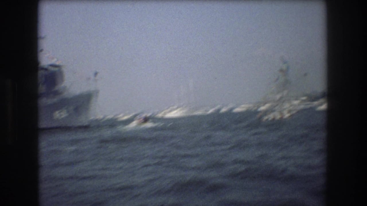 1976 8mm New York Harbor frame with a gray naval ship at left, choppy water, and the Statue of Liberty in the distance.