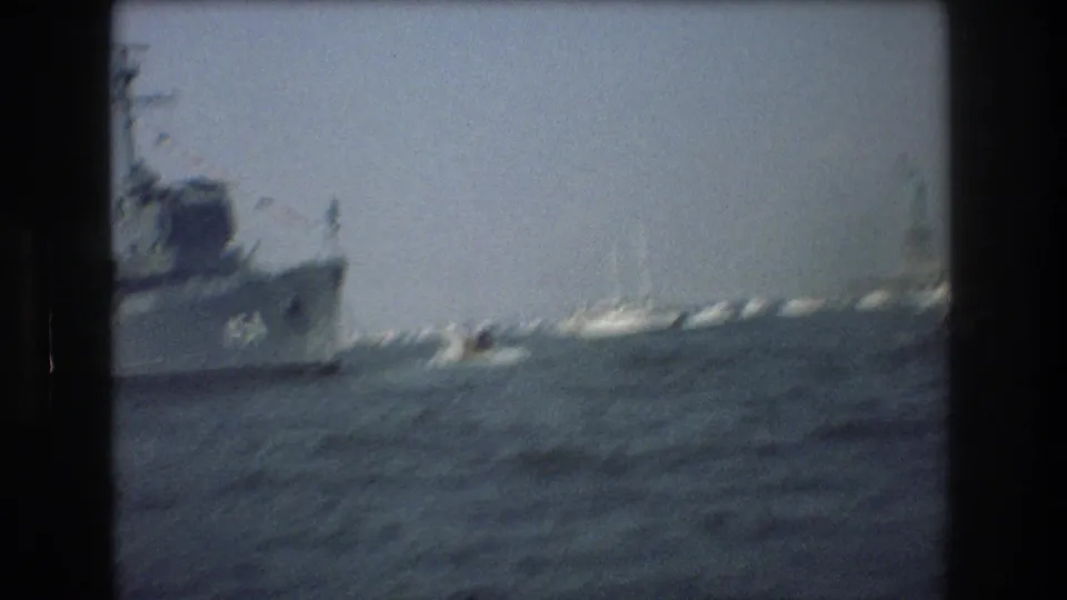 Soft 8mm frame from New York Harbor showing a blurred gray ship at left, open water, and the Statue of Liberty in the distance.