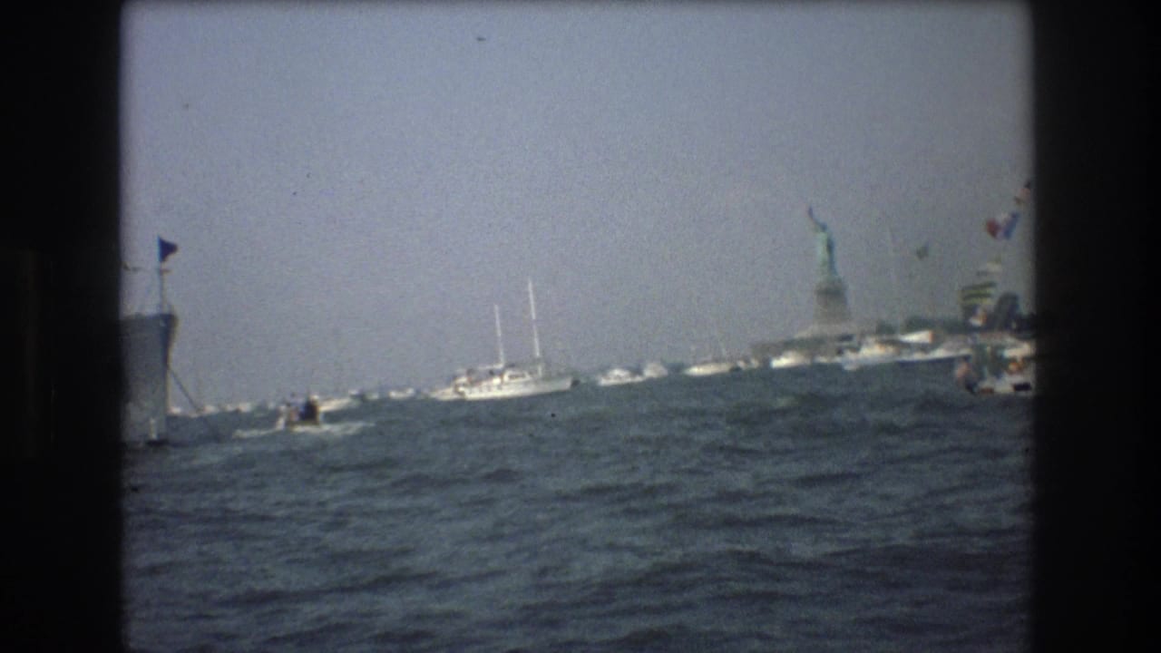 1976 8mm New York Harbor view from the water, with choppy waves, small boats, a larger vessel edge, and the distant Statue of Liberty.