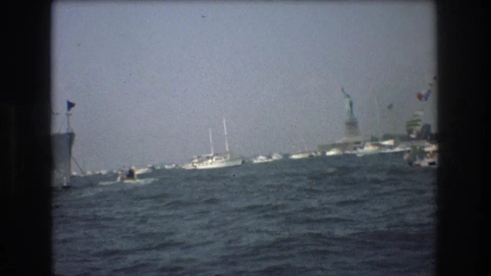 1976 8mm New York Harbor view from the water, with choppy waves, small boats, a larger vessel edge, and the distant Statue of Liberty.