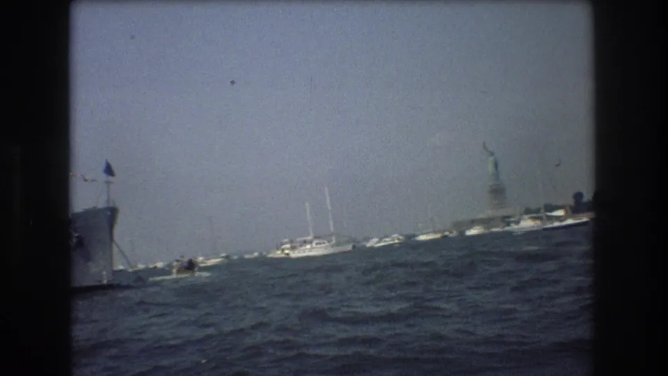 Tilted 1976 home-movie frame of New York Harbor showing choppy water, small craft, a partial vessel at left, and the distant Statue of Liberty.