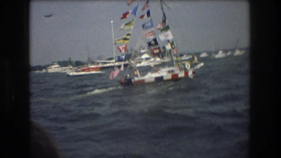 1976 8mm harbor frame showing a sailboat decorated with many flags moving through choppy New York Harbor water among other recreational boats.