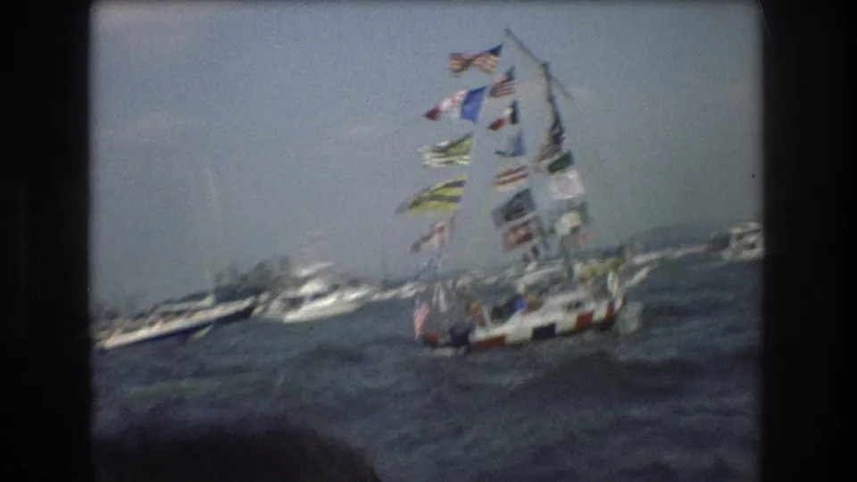 Handheld 1976 New York Harbor view of a sailboat covered with international flags, surrounded by other boats on rough water.