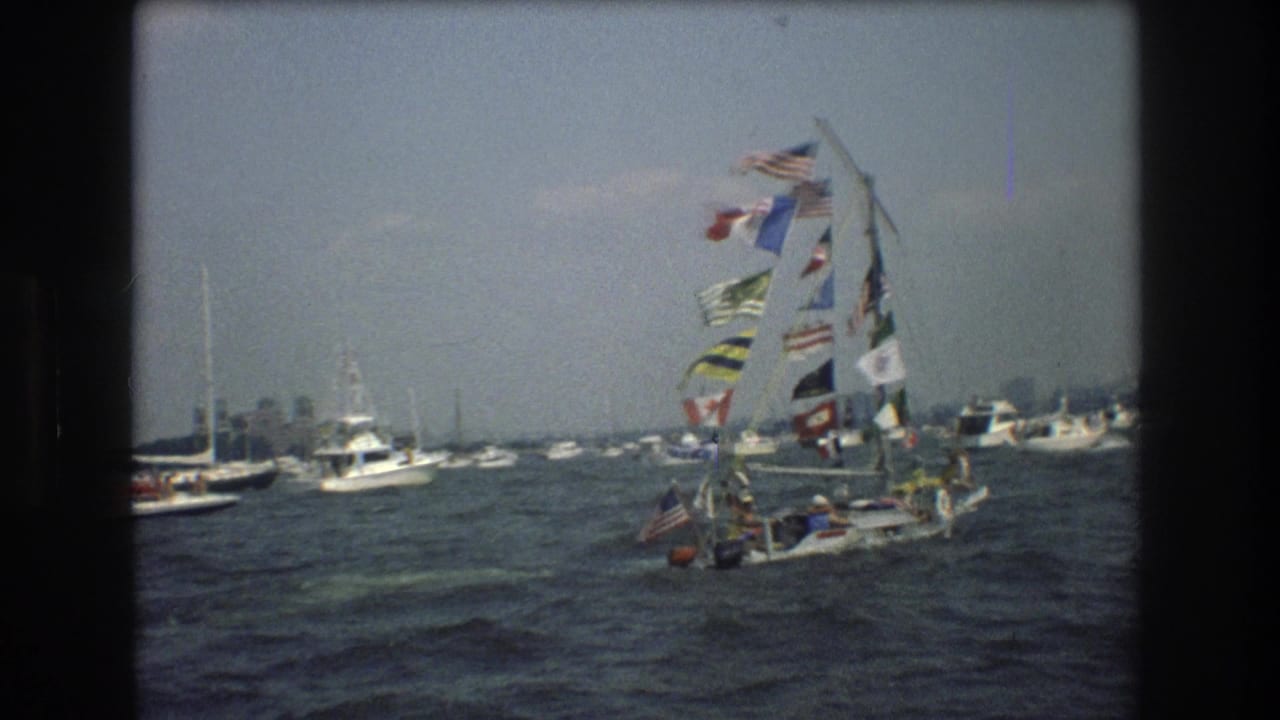 In 1976 New York City harbor footage, a small sailboat decorated with many signal flags passes among other boats on gray, choppy water.