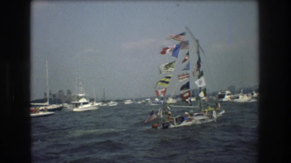 In 1976 New York City harbor footage, a small sailboat decorated with many signal flags passes among other boats on gray, choppy water.