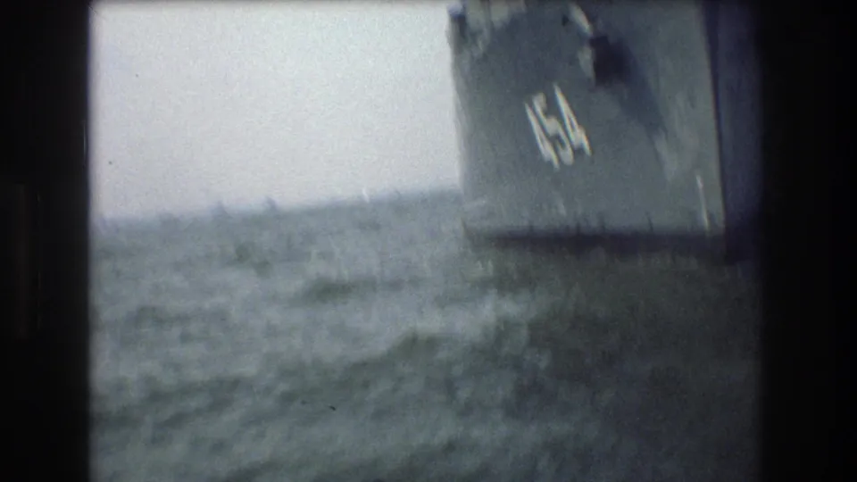 A handheld 8mm frame shows rough harbor water close to the dark side of a United States Navy ship, with part of a white hull number visible.