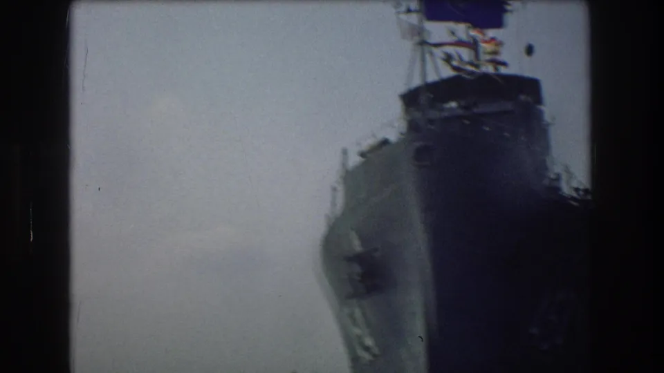 A soft, handheld 8mm view frames the bow and upper structure of a large Navy ship in New York Harbor, with the hull number only partly legible.