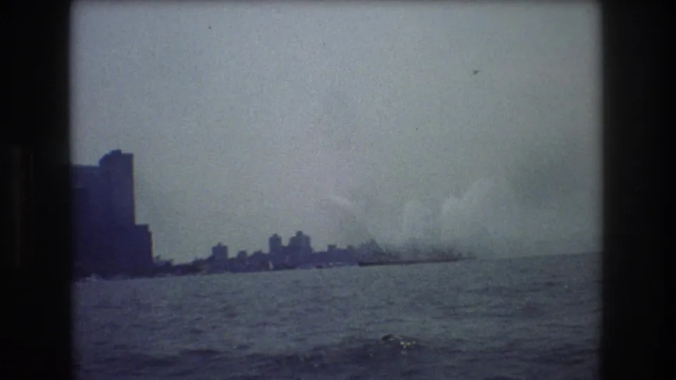 Wide 8mm harbor view across water toward a New York City skyline, with larger buildings at left and smoke or steam near a distant vessel at right.