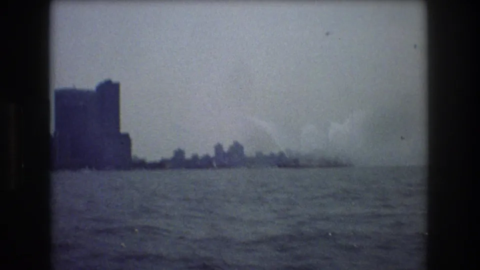 Grainy 8mm view over harbor water toward distant New York City buildings, with smoke or steam rising near a far-off ship or waterfront area.