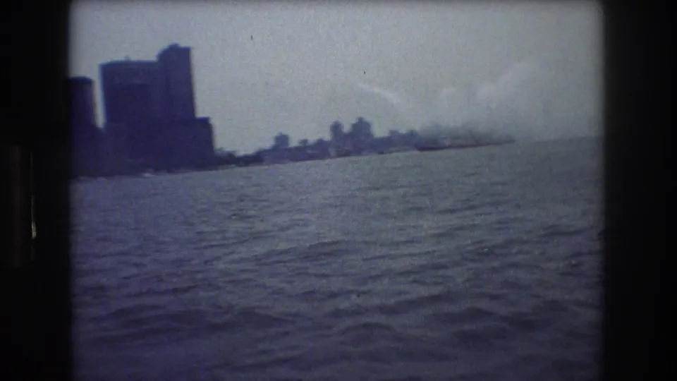 Handheld 8mm harbor shot with rippled water in the foreground, distant New York City skyline, and smoke or steam visible along the far waterfront.
