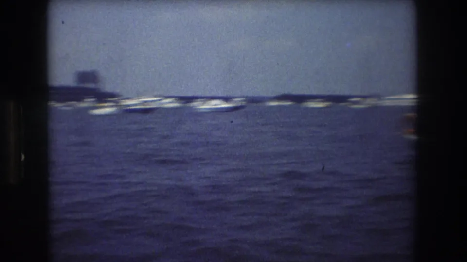 Soft 8mm view of New York City waterfront boating, with distant anchored boats and a low dark shoreline or island shape across the water.