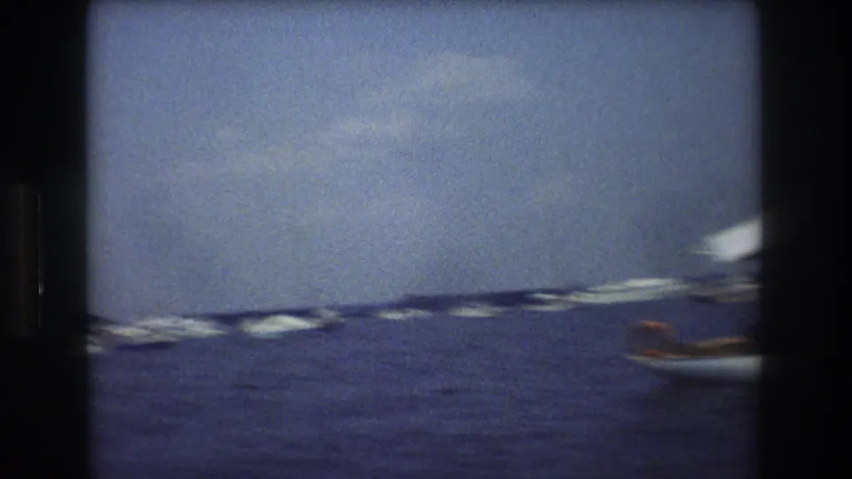Blurred 1976 home-movie frame showing distant boats on the water and a small boat partly entering from the right edge, with a possible seated person visible only indistinctly.