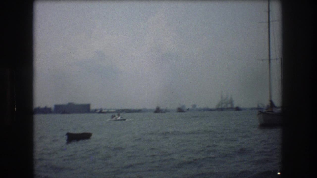 Small boats on harbor water with a distant multi-masted ship and shoreline buildings.