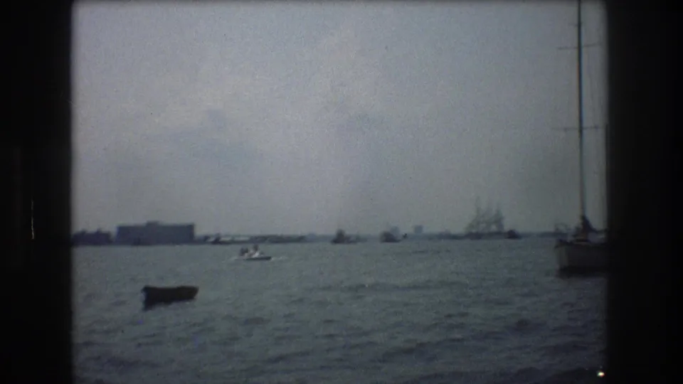 1976 New York City harbor view with a small boat, distant shoreline buildings, and a multi-masted sailing ship visible through grainy 8mm texture.