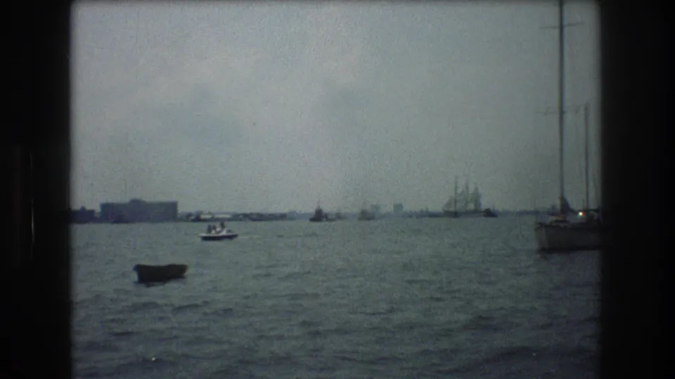 1976 New York City 8mm harbor frame showing a dark small boat in the foreground, a motorboat beyond it, shoreline buildings, and a tall sailboat mast at the right edge.
