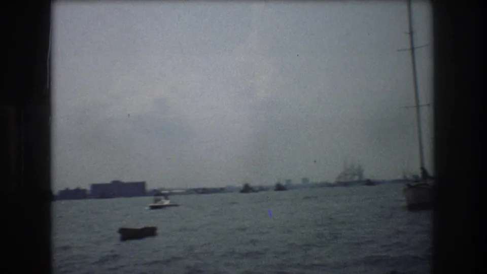 1976 New York City harbor view with choppy water, a dark small boat, a white motorboat, a distant multi-masted ship, and sailboat rigging near the right edge.