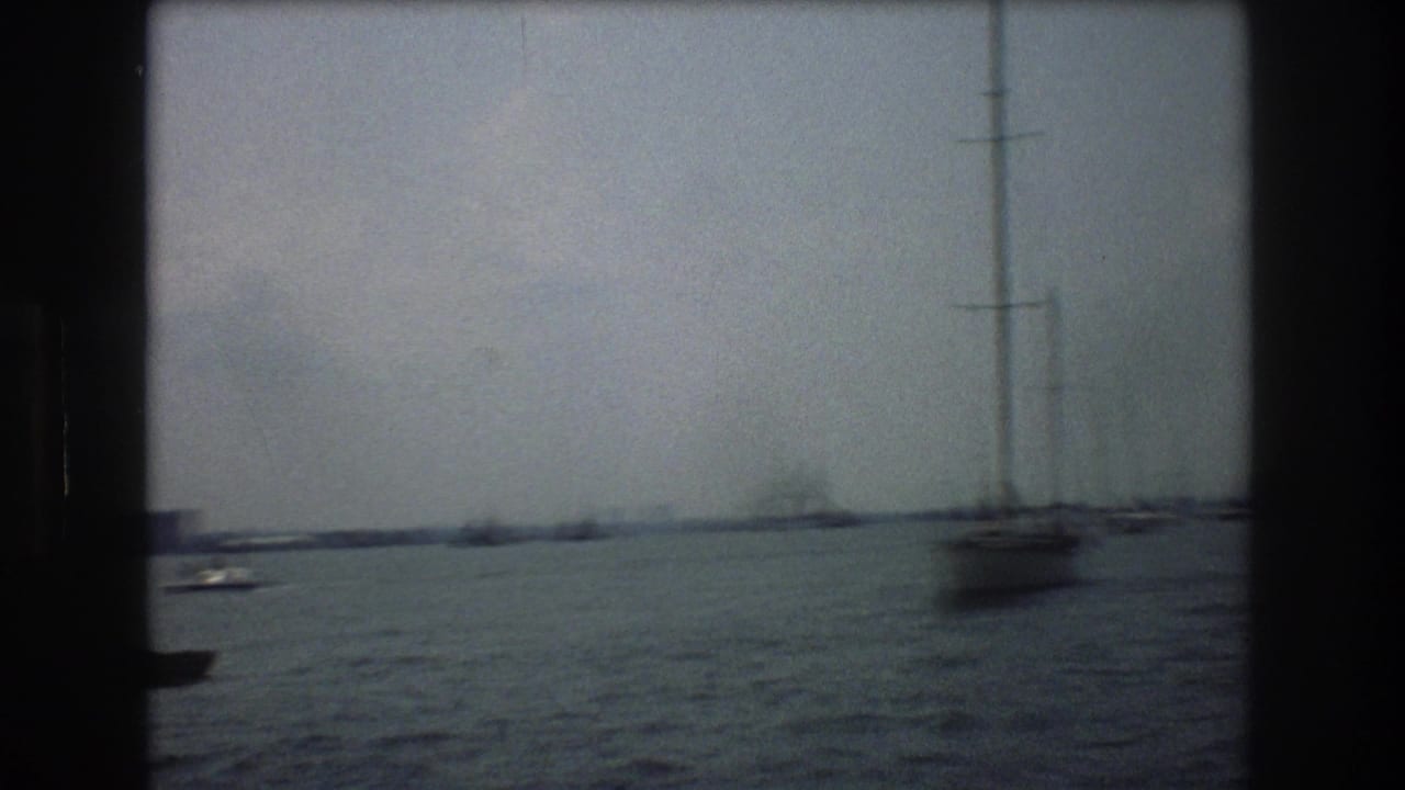 1976 New York City harbor view with choppy water, sailboat masts, distant boats, and a faint shoreline seen through soft 8mm haze.
