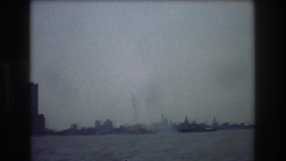 Soft 8mm view across New York City harbor in 1976, showing boats on the water and a dim waterfront skyline beyond them.