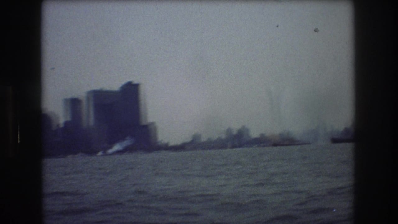 Hazy 1976 New York City skyline seen across open water from a moving boat, with silhouetted buildings and soft blue-gray 8mm grain.