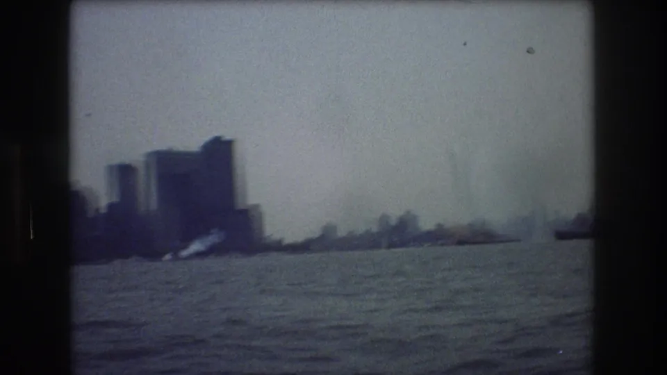 Hazy 1976 New York City skyline seen across open water from a moving boat, with silhouetted buildings and soft blue-gray 8mm grain.