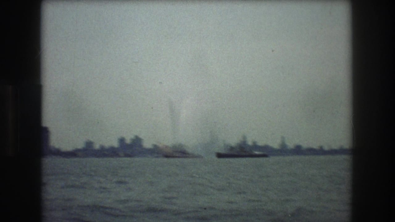 A hazy harbor view toward New York City in 1976, with low distant buildings, small vessels, and open water filmed through soft 8mm exposure.