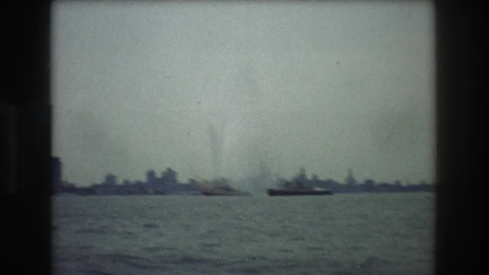 A hazy harbor view toward New York City in 1976, with low distant buildings, small vessels, and open water filmed through soft 8mm exposure.