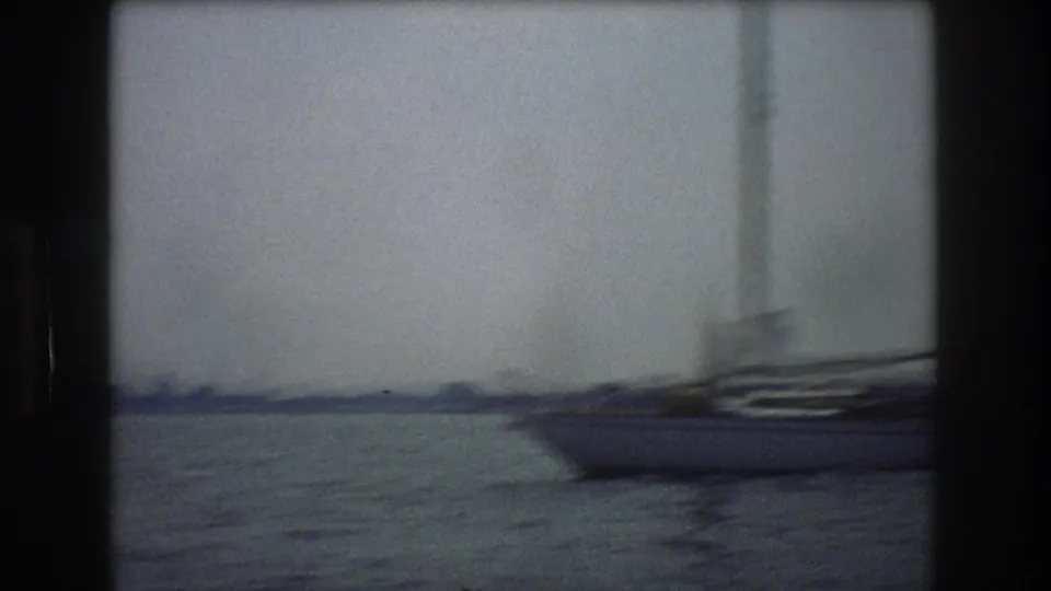 A sailboat passes close to the camera on New York City harbor, blurred by boat motion and soft 8mm focus against a pale skyline distance.