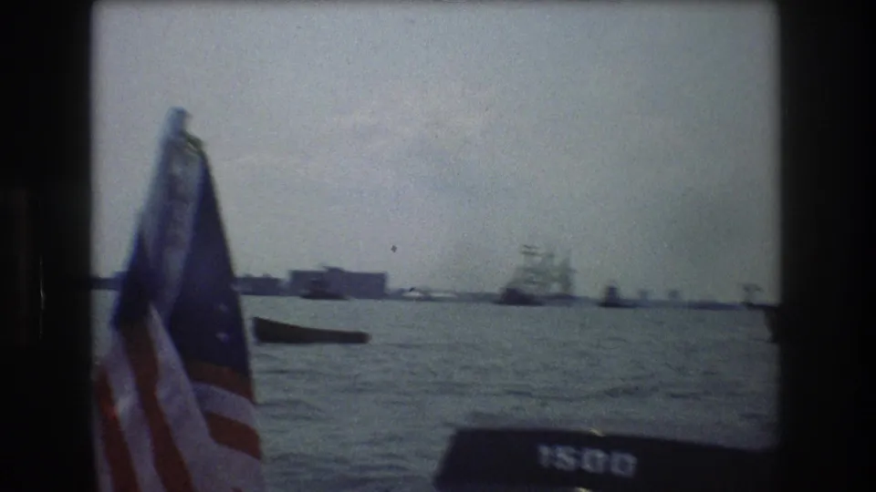 An American flag near the camera frames the 1976 New York City harbor view, with open water, distant boats, and low waterfront structures beyond.