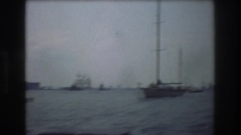 Soft 8mm frame of New York City harbor in 1976, showing sailboats on choppy water with a faint distant shoreline and overcast haze.