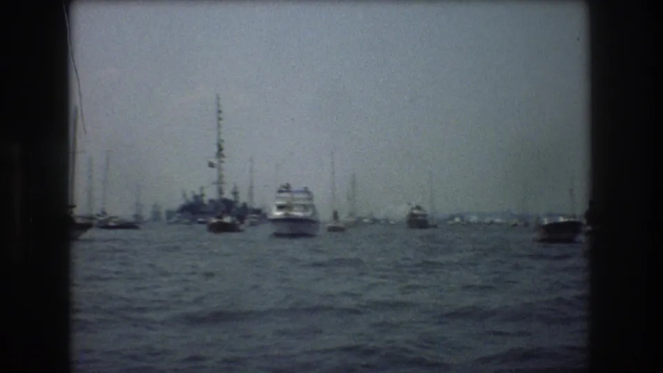 1976 New York City harbor view with sailboats and motor vessels scattered across the water in front of a hazy distant skyline.