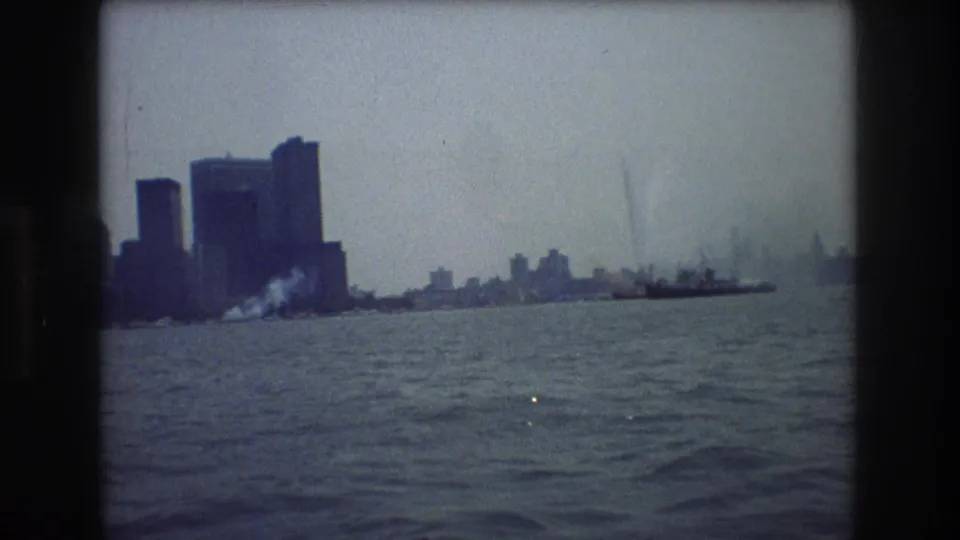 1976 8mm home-movie frame showing a hazy New York City skyline across rippled harbor water from a boat viewpoint.