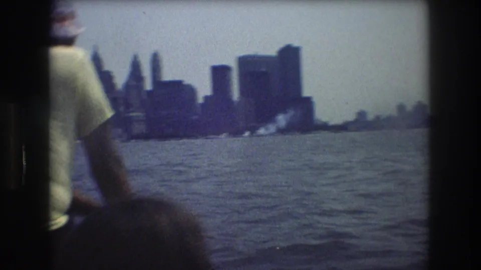 1976 New York City harbor view with a partially visible boat passenger in a light shirt and hat looking toward the skyline.