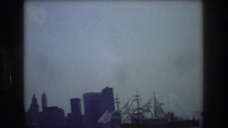 1976 8mm frame showing a distant sailing ship on New York Harbor with the city skyline in soft focus behind it.