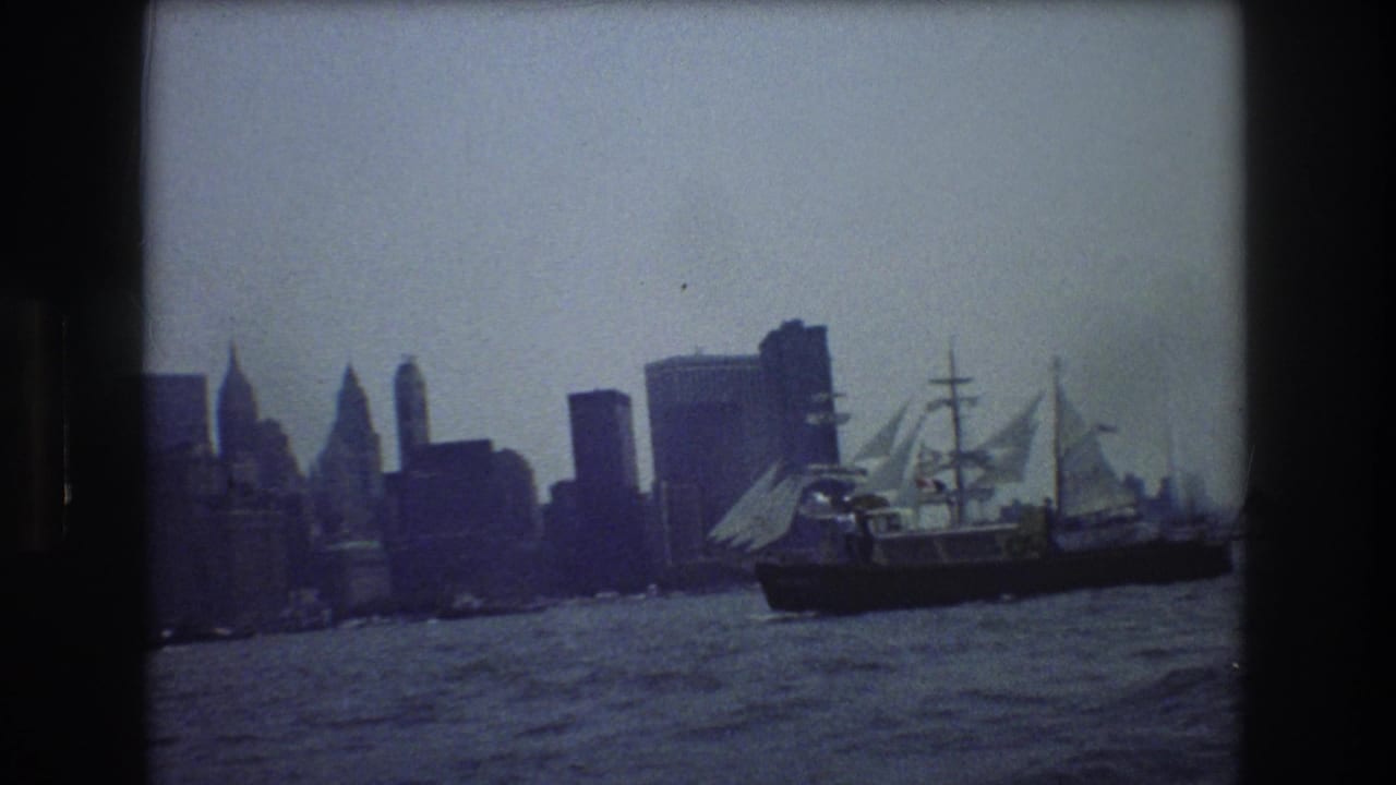 Large multi-masted vessel moves across harbor in front of skyscrapers.