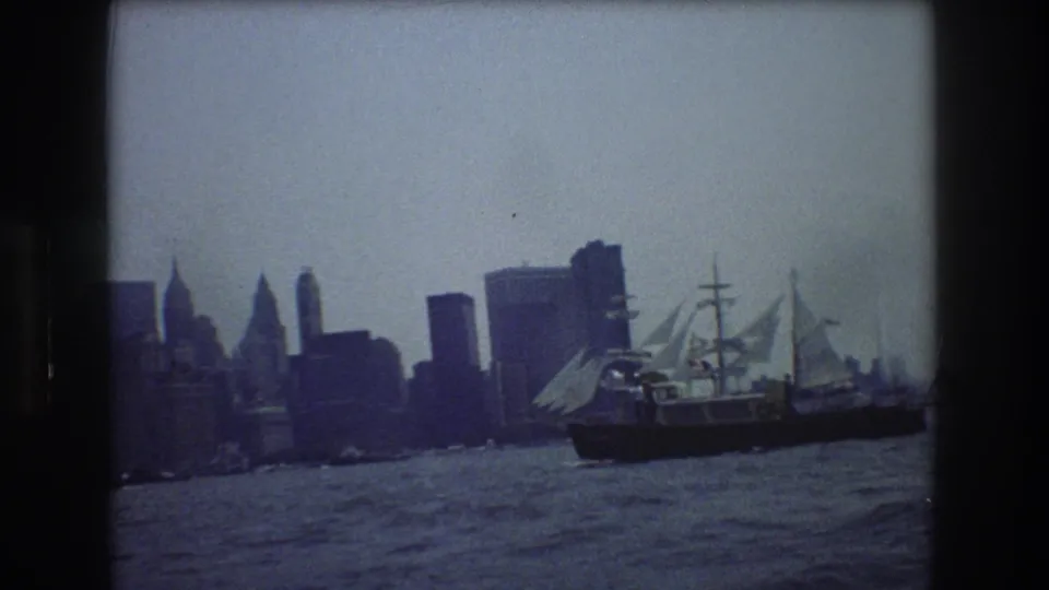 A multi-masted sailing vessel crosses New York Harbor with a hazy Manhattan skyline and choppy water visible in the background.