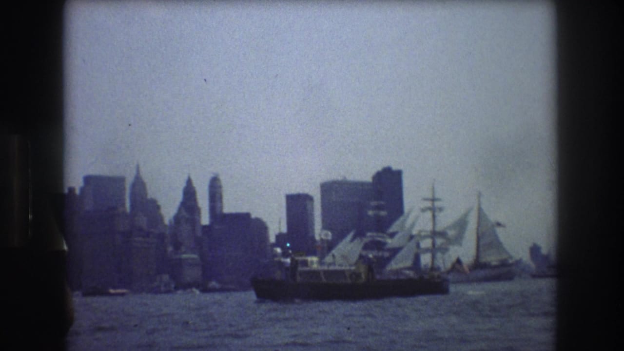 1976 New York City harbor view from a moving boat, with the skyline silhouetted behind a large tall ship on calm water.