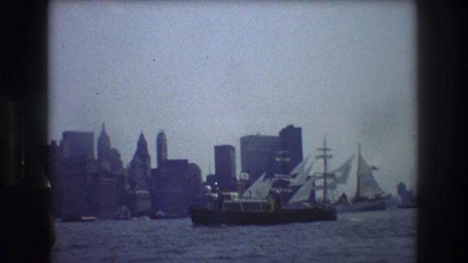 1976 New York City harbor view from a moving boat, with the skyline silhouetted behind a large tall ship on calm water.