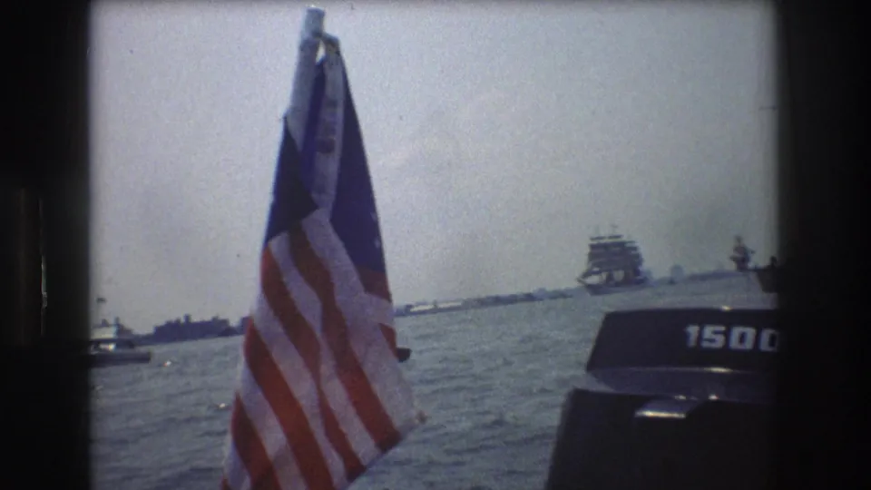 An American flag flutters close to the camera on a boat in New York Harbor, with an outboard motor and distant vessels visible across the water.
