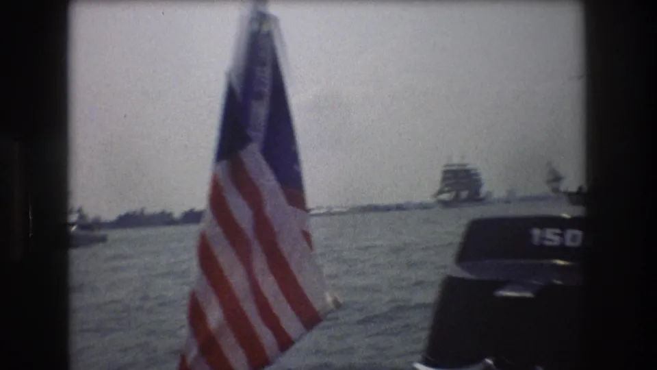 A close foreground American flag and dark outboard motor frame a harbor view, with distant boats and shoreline under a pale sky.