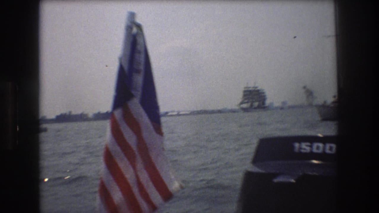 1976 8mm home-movie frame from New York City waters showing an American flag on a boat, choppy harbor water, distant vessels, and part of an outboard motor.