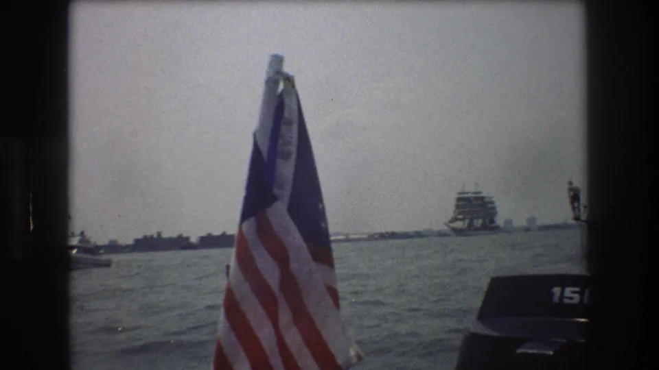 1976 New York City harbor home-movie frame with an American flag in the foreground, open water, a distant vessel, and a partial outboard motor at the right edge.